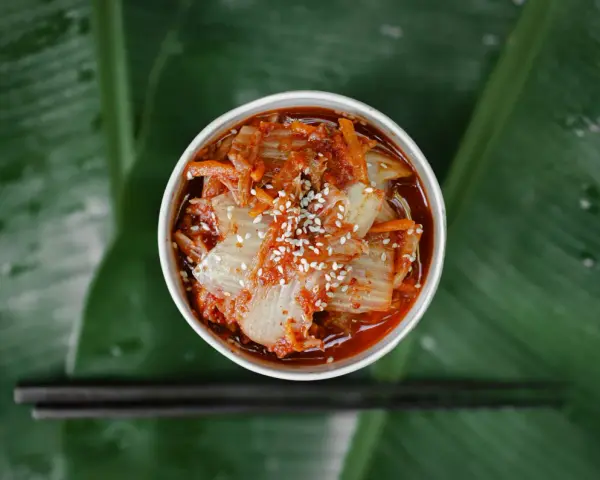 Miseczka z kimchi posypanym sezamem na tle zielonych liści, obok pałeczki.