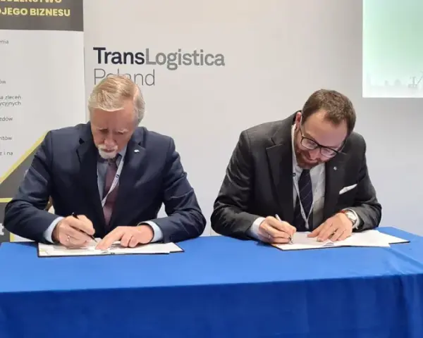 Dwóch mężczyzn w garniturach podpisuje dokumenty. Obok baner z napisem TransLogistica Poland, sugerujący umowę w branży gpw logistics.