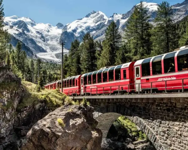 A red train travels across a stone bridge, winding its way from Chur to Tirano through a stunning alpine landscape with snow-capped mountains.