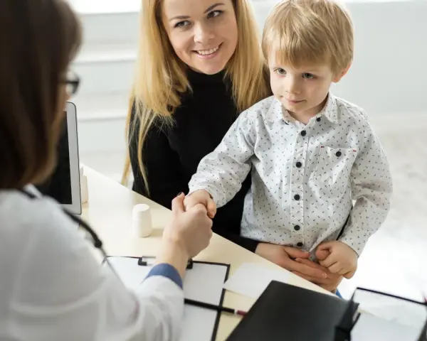 Mama z synkiem u lekarza. Chłopiec ściska dłoń lekarce, a mama uśmiecha się. W tle widać tablet i białą buteleczkę, jakby z lekiem. To ważny moment, być może wpis do książeczki zdrowia.