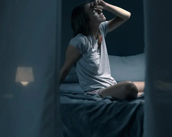 Frau sitzt im Bett, die Stirn gerunzelt, die Hand an der Stirn. Sie fragt sich, wie viel schwitzt man in der Nacht.