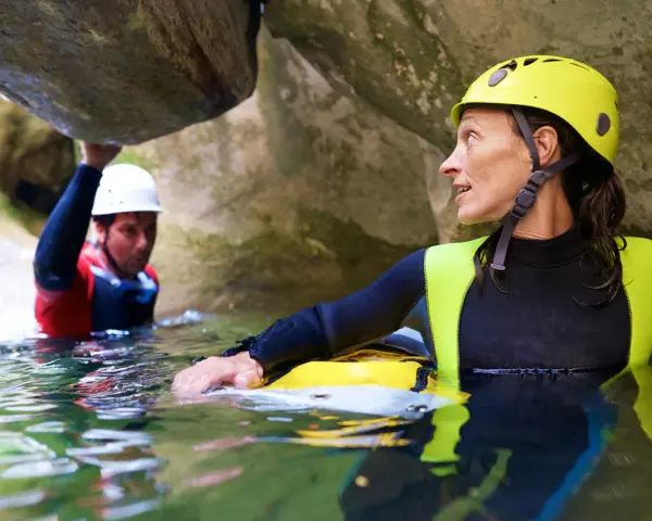 Kobieta w żółtym kasku i piankowym stroju zanurzona w wodzie, obok mężczyzna w białym kasku. To właśnie canyoning – przygoda w kanionie.