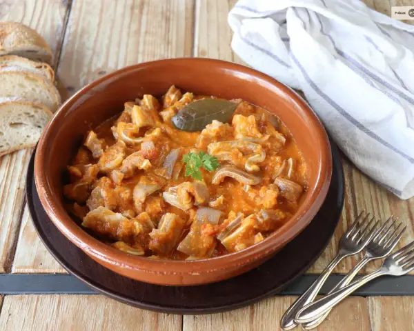 Guiso casero de oreja de cerdo en salsa de tomate, servido en cazuela de barro, acompañado de pan rústico y tenedores.