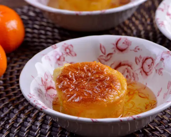 Un esponjoso baba al ron bañado en almíbar, adornado con ralladura de naranja, en un cuenco floral. Cerca, mandarinas frescas.