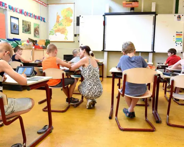 Dzieci siedzą w klasie, ucząc się. Nauczycielka pochyla się nad jednym z uczniów. Ile jest klas w Anglii? Tego nie wiemy, ale ta lekcja wygląda na interesującą.