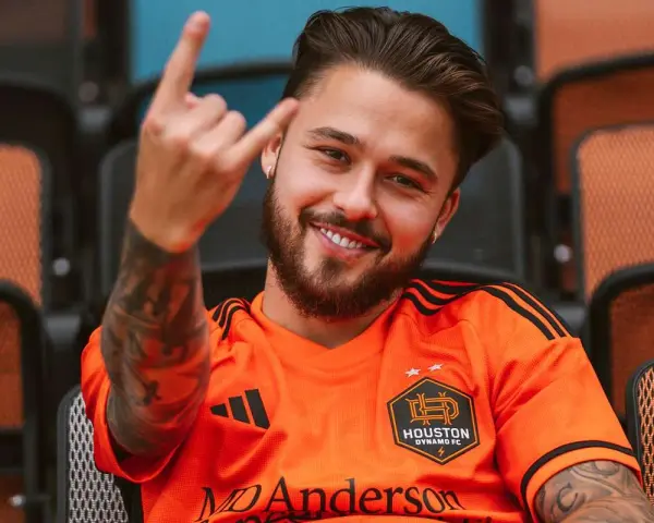 Uśmiechnięty Adam Kowalczyk w koszulce Houston Dynamo FC pozdrawia gestem rock and rolla, siedząc na trybunach.