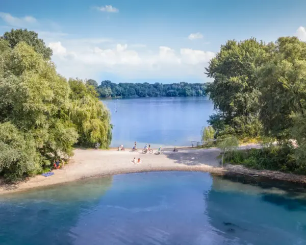 Sommerliche Idylle am Strandbad Wedau: Menschen genießen den Tag am Seeufer, einige radeln, andere entspannen im Sand.