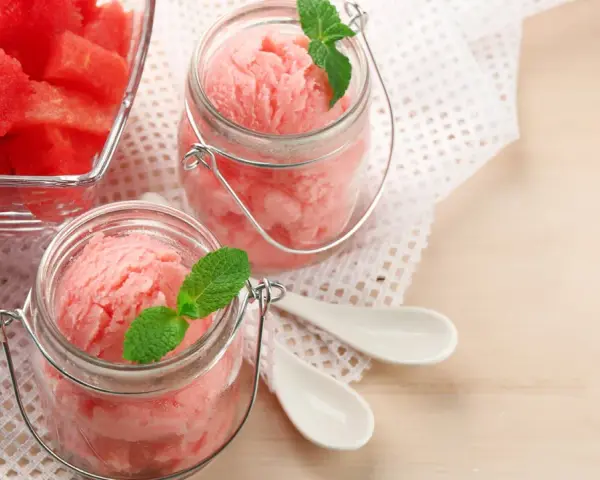 Erfrischendes Wassermelonen-Eis in Gläsern, garniert mit Minze. Daneben ein Schälchen mit frischen Wassermelonenstücken.