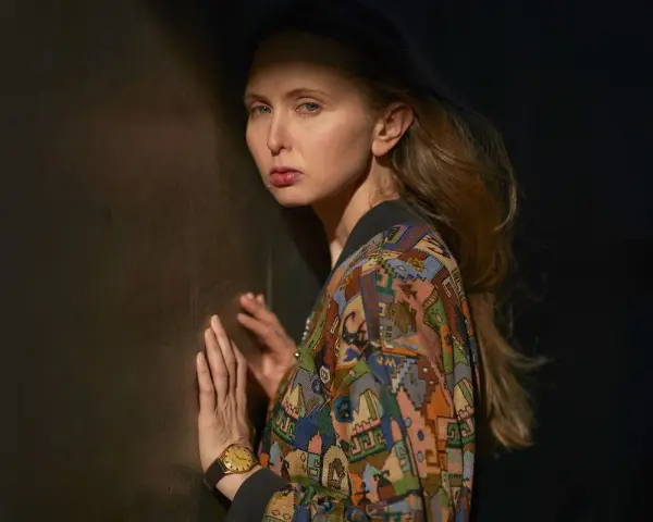 Justyna Wasilewska w kolorowej kurtce opiera się o ciemną ścianę, jej twarz oświetla cień.