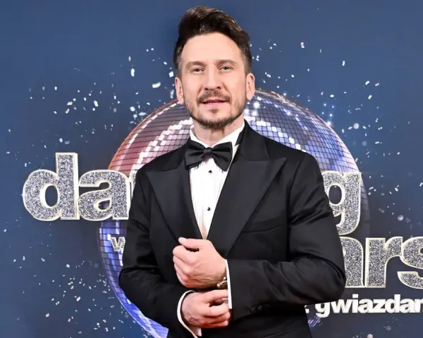 Tomasz Barański w czarnym smokingu pozuje na tle błyszczącej kuli dyskotekowej i napisu "Dancing with the Stars".