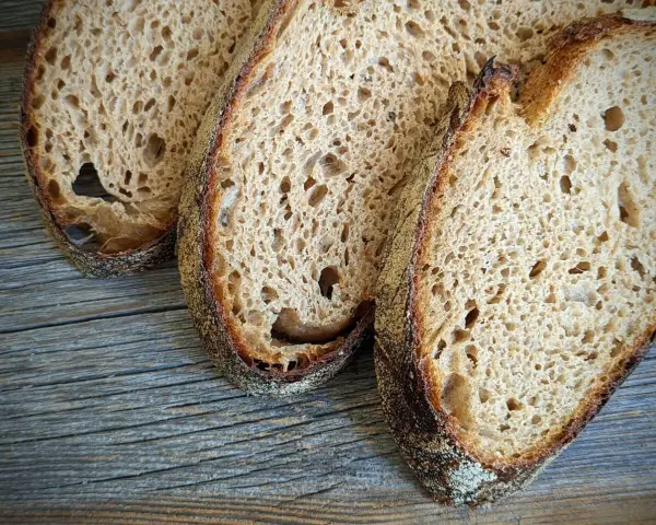 Drei Scheiben rustikales Sauerteigbrot liegen auf Holz. Der Sauerteig ist zu sauer, aber die Kruste ist perfekt gebräunt.