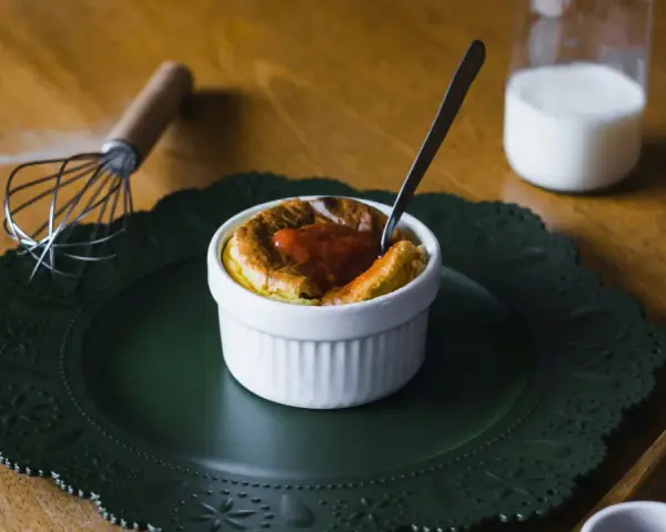 Delicioso souffle de vainilla con salsa de fresa, listo para disfrutar. A su lado, un batidor y un vaso de leche.