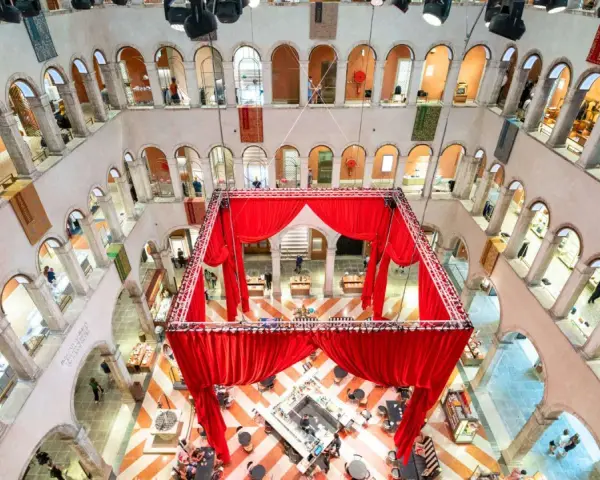 Interni del Fondaco dei Tedeschi con un'installazione di tende rosse al centro, che incornicia un'area bar e tavoli. Le arcate dei piani superiori ospitano negozi.