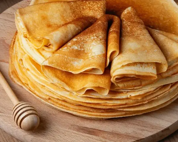 Pile de crêpes dorées, prêtes à être garnies. Une cuillère à miel en bois repose à côté, suggérant une délicieuse recette de crêpes.