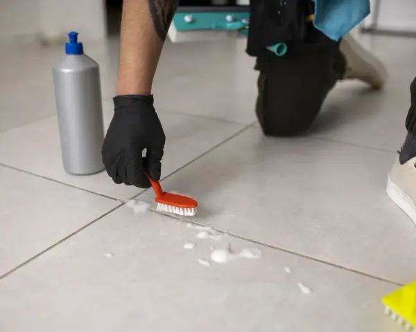 Osoba w czarnej rękawiczce czyści podłogę z płytek małą szczoteczką i pianą.