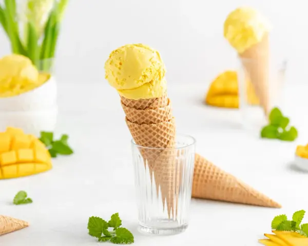 Zwei Kugeln cremiges Mango Eis in Waffelhörnchen, umgeben von frischen Mangostücken und Minze. Ein süßer Genuss!