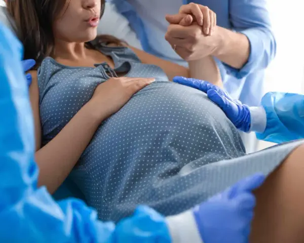 Donna in travaglio, mani che si stringono, personale sanitario con guanti blu. Si percepisce la tensione e l'attesa per lo scollamento membrane.
