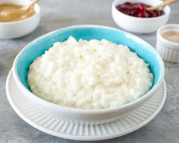 Cremiger Milchreis im blauen Schälchen, daneben Apfelmus, Kirschen und Zimt. Perfekt für ein einfaches Milchreis Rezept.