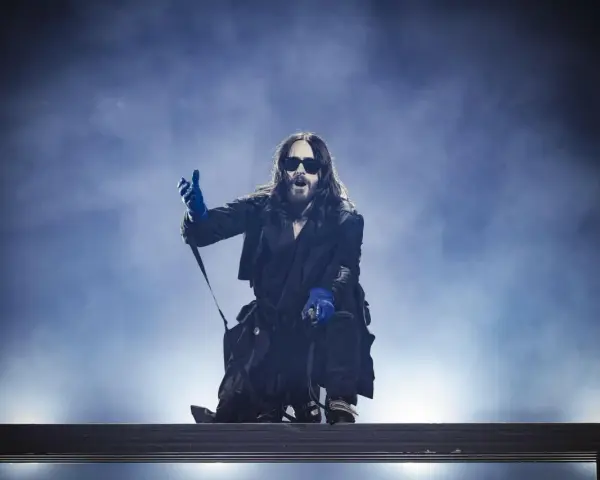 Jared Leto w niebieskich rękawiczkach, w okularach i długim płaszczu, klęczy na scenie. Jego zespół pewnie czeka na jego znak.
