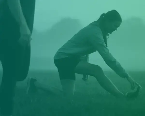 Två personer stretchar utomhus i dimma. En kvinna på knä sträcker sig mot sin fot. Detta kan vara en del av rehab benhinneinflammation.