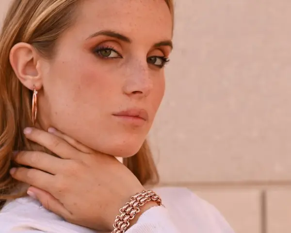 Donna con lentiggini e gioielli in oro rosa, un bracciale a maglie larghe e orecchini a cerchio.