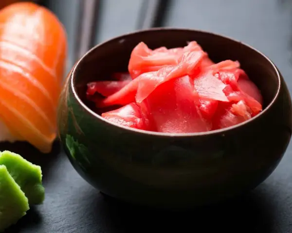 Miseczka z różowym imbirem do sushi, obok kawałek łososia i zielone wasabi.