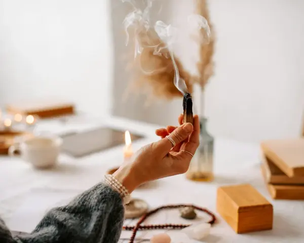 Mano sosteniendo un palo santo humeante, rodeada de velas, libros y cristales, creando un ambiente de sanación y paz.