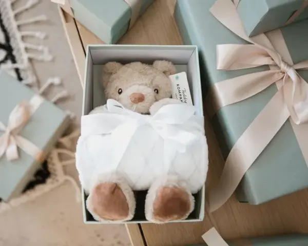 En mjuk nallebjörn i en presentbox, omgiven av fler presenter med ljusrosa band. En perfekt välkommen till världen present.