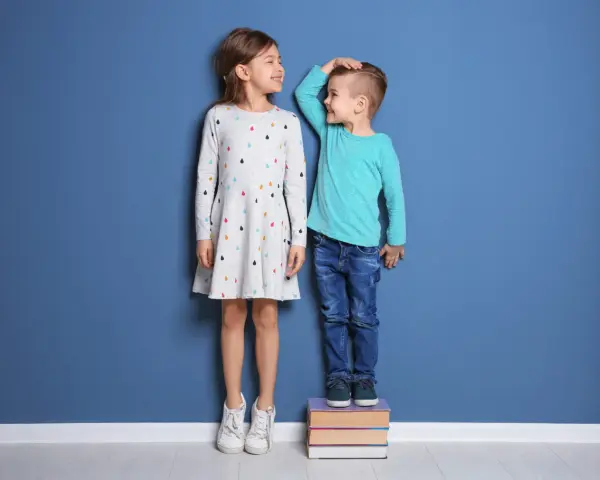 Dziewczynka i chłopiec mierzą się na tle niebieskiej ściany. Chłopiec stoi na książkach, by dorównać dziewczynce. Wiedza i zdrowie to klucz do tego, jak wspomóc wzrost dziecka.