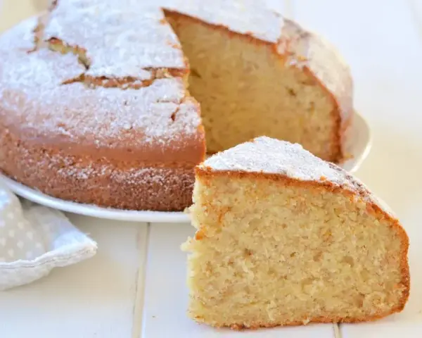Una fetta di soffice torta di banane mature, spolverata di zucchero a velo, accanto alla torta intera su un piatto bianco.