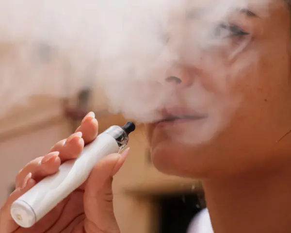 Una mujer exhala vapor de un vaper blanco. El vapor parece rodear su rostro, sugiriendo que el vaper encharca los pulmones.