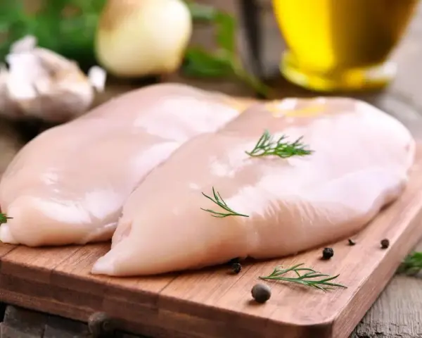 Dwie surowe piersi z kurczaka na desce, obok czosnek, cebula i koperek. Zastanawiasz się, ile białka ma pierś z kurczaka?