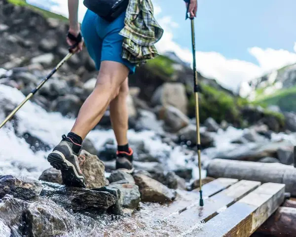 Une personne en tenue de ppg sport traverse un ruisseau sur un pont de bois, bâtons de randonnée en main. Paysage de montagne.