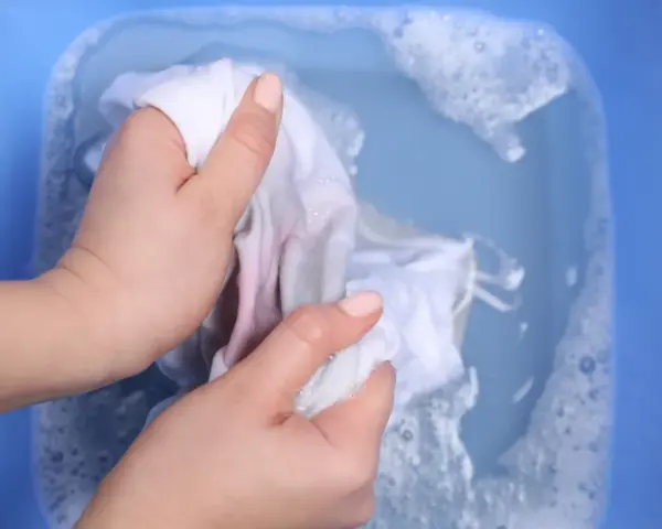 Ręce piorą białą tkaninę z delikatnymi plamami z wiśni w niebieskiej misce z wodą i pianą.