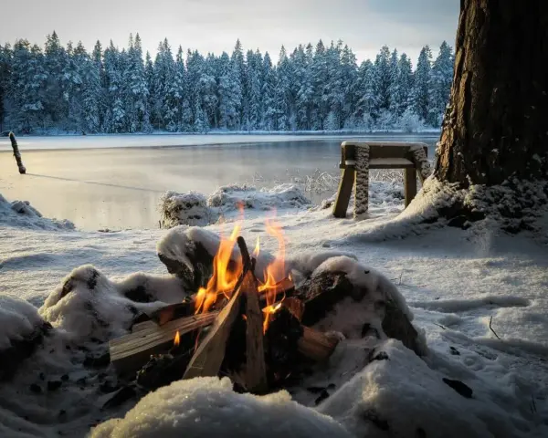 Ein Lagerfeuer wärmt beim Zelten im Winter am gefrorenen See. Schnee bedeckt die Ufer und den Wald im Hintergrund.