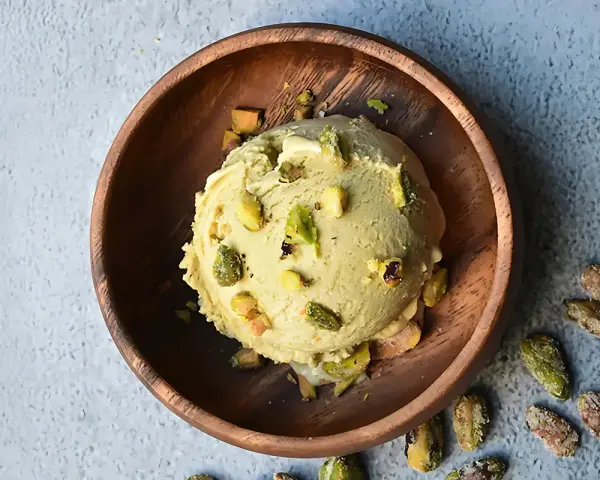 Cremoso helado de pistacho servido en un cuenco de madera, adornado con trozos de pistacho y acompañado de más pistachos enteros esparcidos alrededor.
