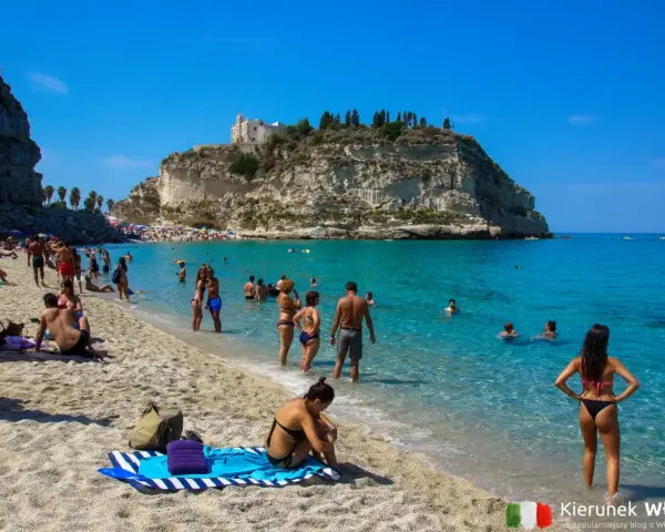 Słoneczna plaża z turkusową wodą i klifem, na którym stoi kościół. Ludzie kąpią się i opalają, ciesząc się jednymi z najpiękniejszych plaż we Włoszech.