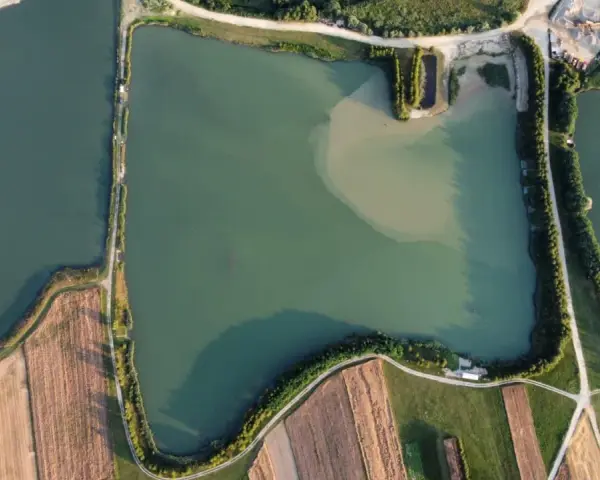 Widok z lotu ptaka na zielone oczko wodne, otoczone polami i drogami. Idealne miejsce na spokojne łowisko.