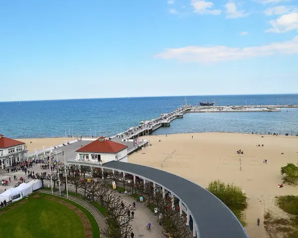 Sopot beach z molo, kawiarniami i spacerującymi ludźmi. W oddali widać statki.
