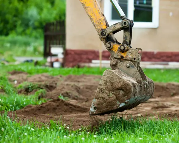 Błotnista łyżka koparki wykonuje niwelację terenu wokół domu.