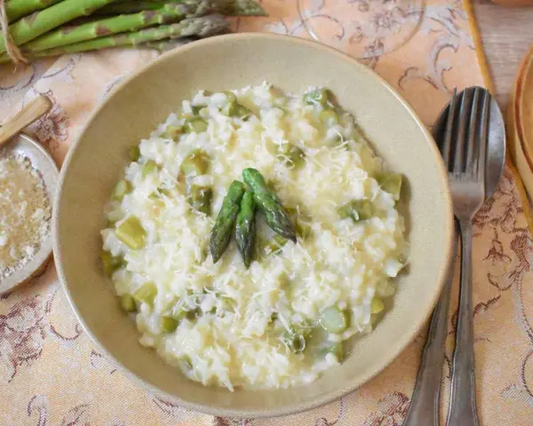 Kremowe risotto ze szparagami, posypane parmezanem, z trzema zielonymi szparagami na wierzchu. Obok miseczka z tartym serem i pęczek świeżych szparagów.
