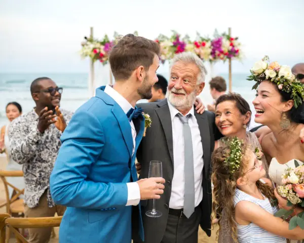 Szczęśliwy ojciec pana młodego w eleganckim garniturze śmieje się z synem na plażowym weselu.