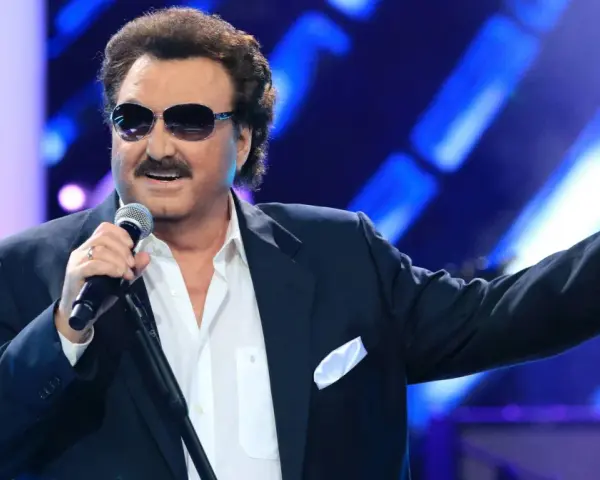 Krzysztof Krawczyk w okularach przeciwsłonecznych i granatowej marynarce śpiewa na scenie, gestykulując prawą ręką.
