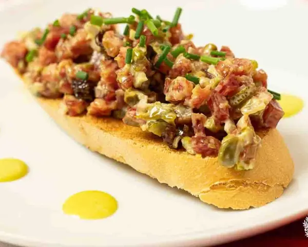 Tosta con steak tartar de salchichón y cebollino picado, decorada con puntos de salsa amarilla.