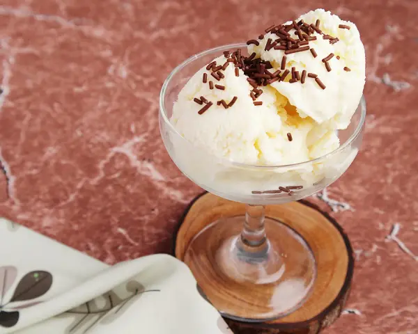 Cremiges Litschi Eis mit Schokostreuseln in einem Glas auf einem Holzuntersetzer.