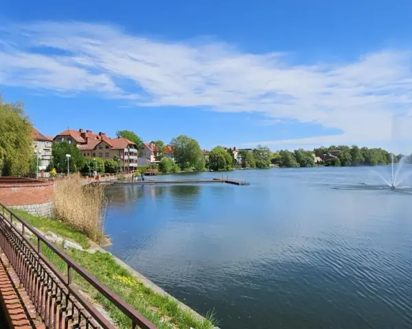Spokojne jezioro zamkowe z fontanną i drewnianym pomostem, otoczone zielenią i zabudowaniami.