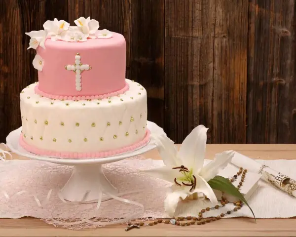 Elegancki dwupoziomowy tort na chrzest w różowo-białych barwach, ozdobiony krzyżem i kwiatami. Obok lilia, różaniec i świeca.