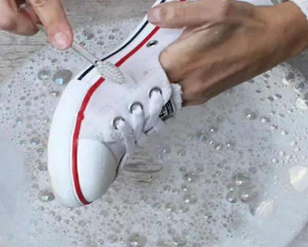 Manos limpian zapatillas de tela de color con cepillo en agua con jabón.
