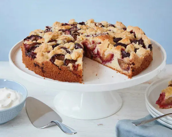 Ein einfacher Pflaumenkuchen mit Streuseln auf einem Tortenständer, daneben eine Schüssel Schlagsahne und ein Tortenheber.