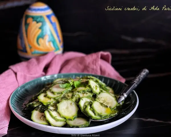 Zucchine marinate cotte, condite con erbe aromatiche, servite su un piatto verde. Sullo sfondo, un oggetto decorativo colorato e un tovagliolo rosa.